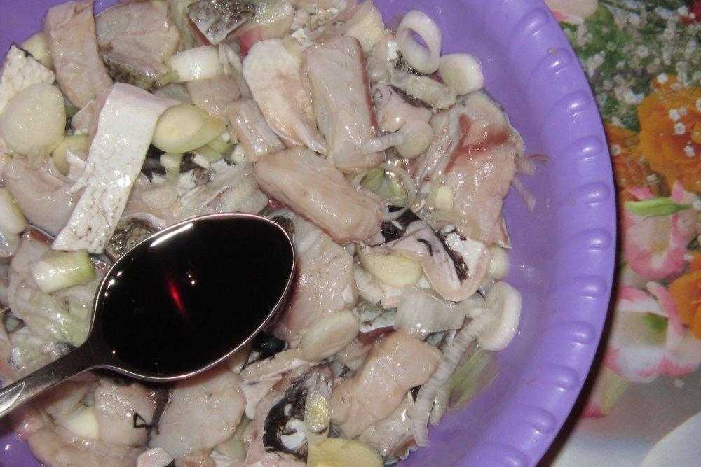 в большой фиолетовой тарелке лежит толстолобик, нарезанный небольшими кусочками, репчатый лук, нарезанный кольцами, соевый соус в столовой ложке добавляют в тарелку с рыбой